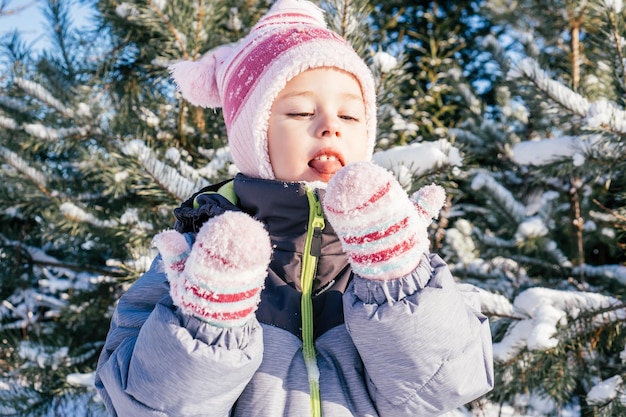 Petite Fille De 34 Ans En Salopette D'hiver Chapeau Et Mitaines Se Dresse Contre Les Pins Et Les Sapins Enneigés Mange De La Neige