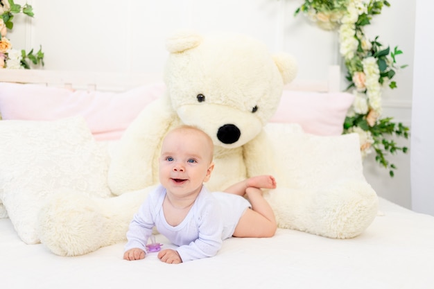 Une petite fille de 6 mois est allongée sur un lit blanc à la maison avec un grand ours en peluche