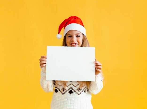 Une petite fille de 8 ans aux cheveux blonds coiffée d'un chapeau de Père Noël tient une feuille de papier blanc sur fond jaune.