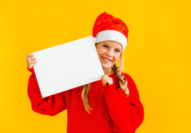 Une petite fille de 8 ans aux cheveux blonds coiffée d'un chapeau de Père Noël tient une feuille de papier blanc sur fond jaune.