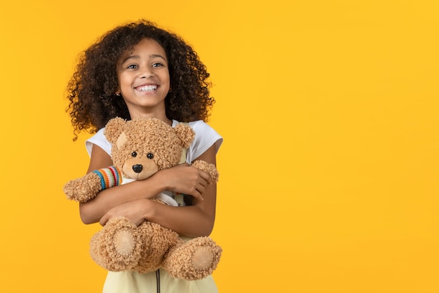 Petite fille africaine mignonne étreignant le jouet d'ours en peluche isolé sur fond jaune