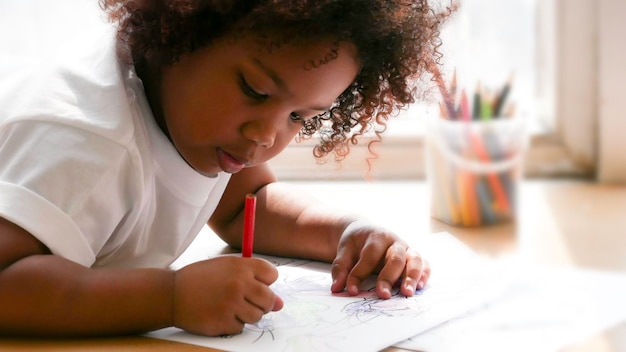 Petite fille africaine peignant et dessinant sur du papier. Fille d'âge préscolaire allongée à écrire sur le sol.