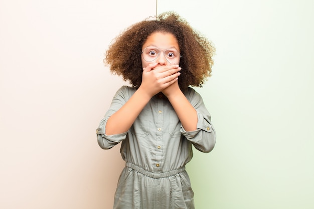 Petite fille afro-américaine coning bouche avec les mains avec une expression choquée et surprise, gardant un secret ou disant oops sur un mur plat