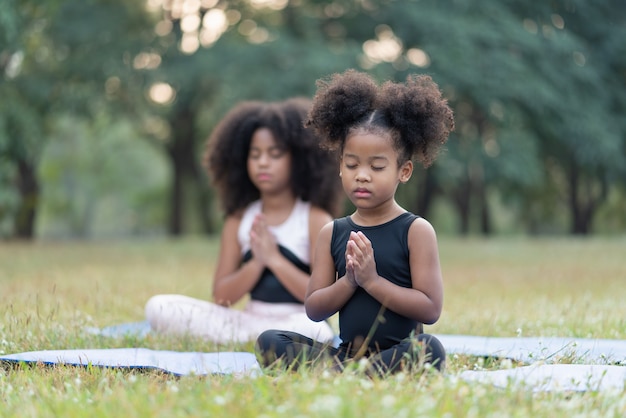 Petite fille afro-américaine souriante et assise sur un tapis roulant, pratique le yoga de méditation dans un parc en plein air