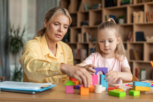 Photo petite fille d'âge préscolaire jouant avec des blocs de bois et aide l'éducateur enseignant assis à table au bureau