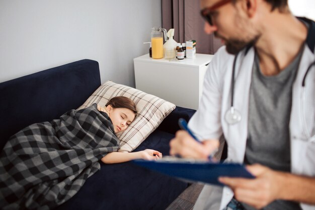 Petite fille allongée sur le lit et dormant. Elle a couvert de couverture. Jeune médecin de sexe masculin la regarde et écrit.