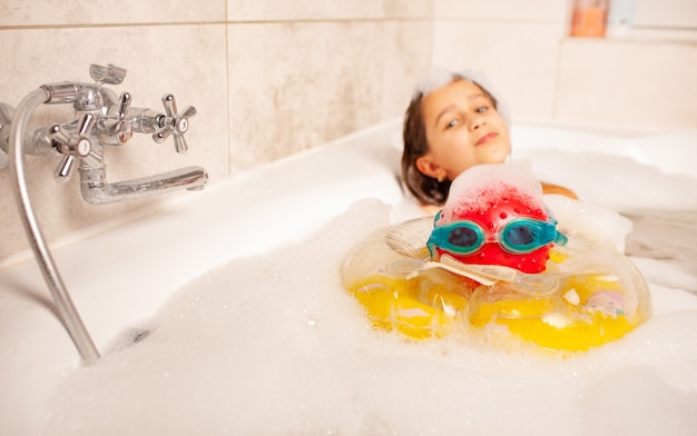 Une petite fille amusante se baigne dans un bain avec de la mousse et joue au ballon et à des lunettes de natation. Concept de loisirs pour enfants à la maison. Espace de copie