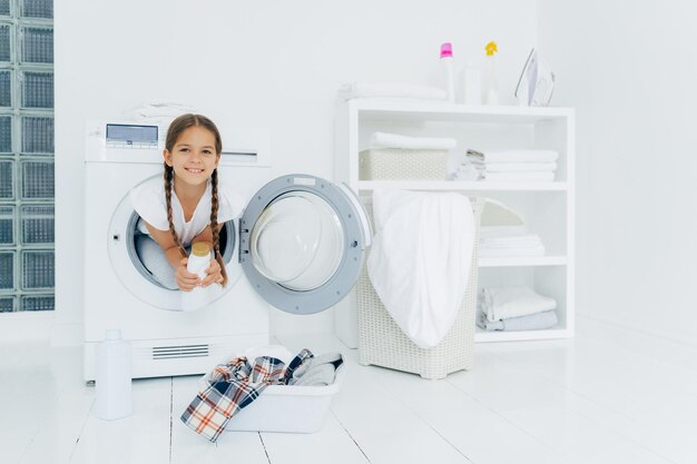 Une petite fille à l'apparence attrayante s'amuse et pose à l'intérieur de la machine à laver contient du détergent se prépare pour le lavabo avec des vêtements à mettre dans la machine à laver sur un sol blanc Jour de lessive à la maison