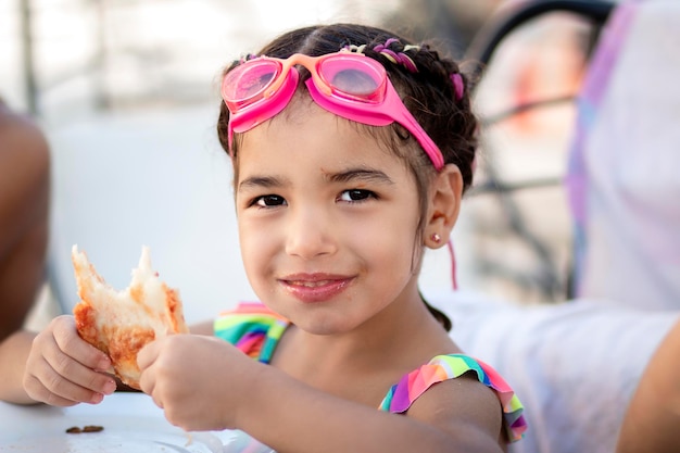 Petite fille appréciant de manger après être sortie de la piscine avec des lunettes de natation sur la tête.