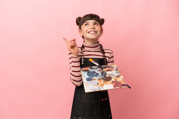 Petite fille artiste tenant une palette isolée sur un mur rose pointant vers le haut une excellente idée