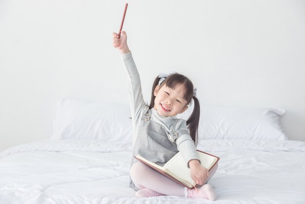 Petite fille asiatique assise sur le lit et sourire tout en tenant le crayon et le cahier