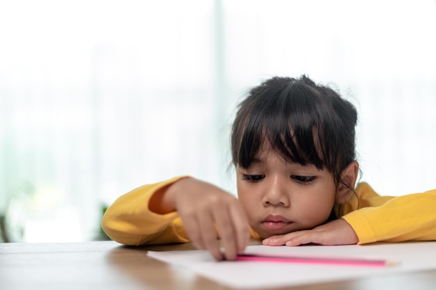 Petite fille asiatique assise seule et regardant dehors avec un visage ennuyé Enfant d'âge préscolaire allongé sur la table avec un enfant gâté triste ennuyé par les devoirs