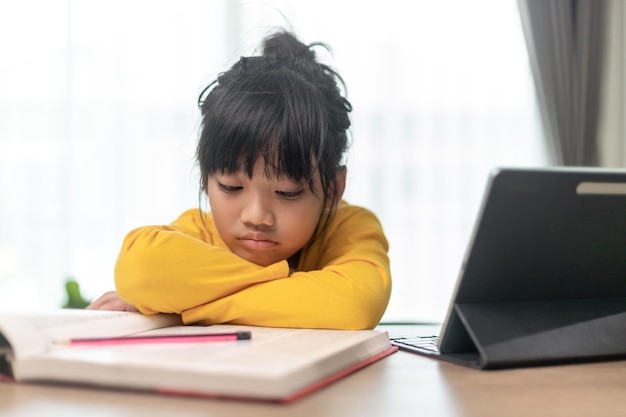 Petite fille asiatique assise seule et regardant dehors avec un visage ennuyé Enfant d'âge préscolaire allongé sur la table avec un enfant gâté triste ennuyé par les devoirs