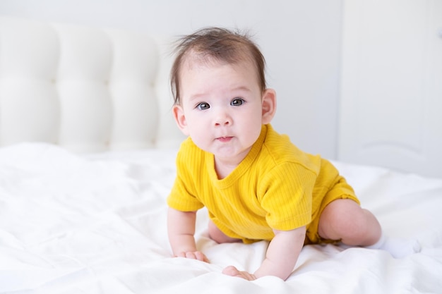 Une petite fille asiatique en bonne santé de 5 mois en body jaune sur le lit sur une literie blanche