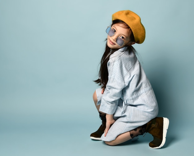 Petite fille asiatique brune à lunettes de soleil, robe chemise surdimensionnée, béret marron, bottes. Elle souriante, posant assise sur le côté sur fond bleu. Enfance, mode, style hipster. Gros plan, espace de copie