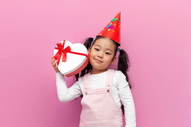 Une petite fille asiatique célèbre son anniversaire dans une casquette festive et tient un coeur cadeau sur fond rose