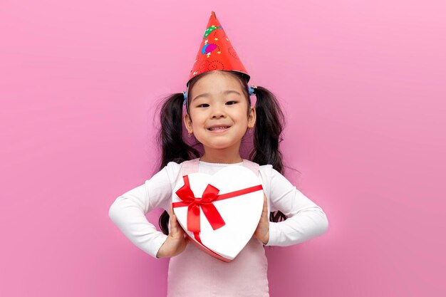 Une petite fille asiatique célèbre son anniversaire dans une casquette festive et tient un coeur cadeau sur fond rose