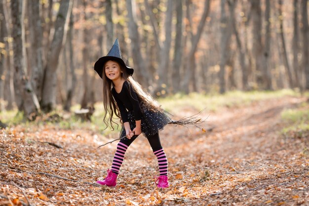 Une petite fille asiatique en costume de sorcière pour Halloween se promène dans la forêt avec un seau pour les bonbons