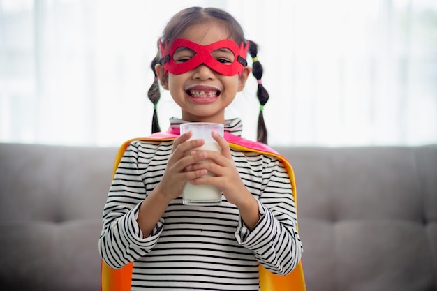 Une petite fille asiatique en costume de super-héros buvant du lait à la maison.