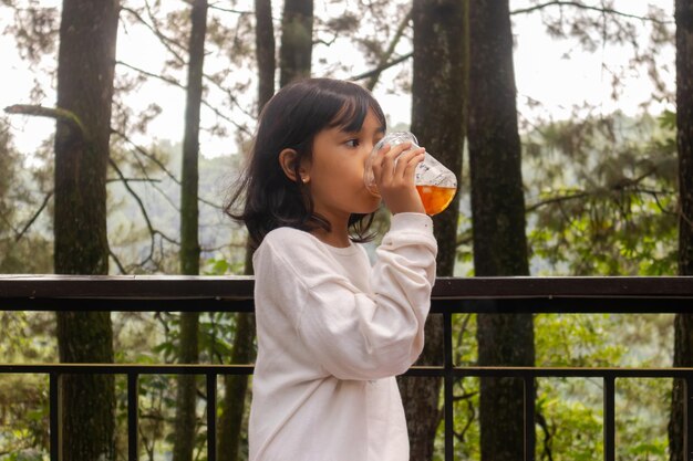 Une petite fille asiatique debout et buvant une tasse en plastique de thé glacé avec la nature ou les arbres en arrière-plan