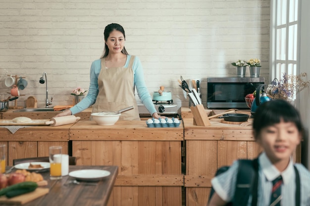 Petite fille asiatique élève du primaire en uniforme avec sac à dos quittant la maison pour étudier après avoir dit au revoir à sa mère. femme au foyer élégante reste dans la cuisine en souriant en regardant sa petite fille