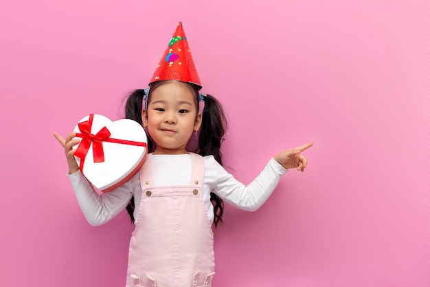 petite fille asiatique fête son anniversaire en casquette de fête et tient un coeur cadeau sur fond rose isolé