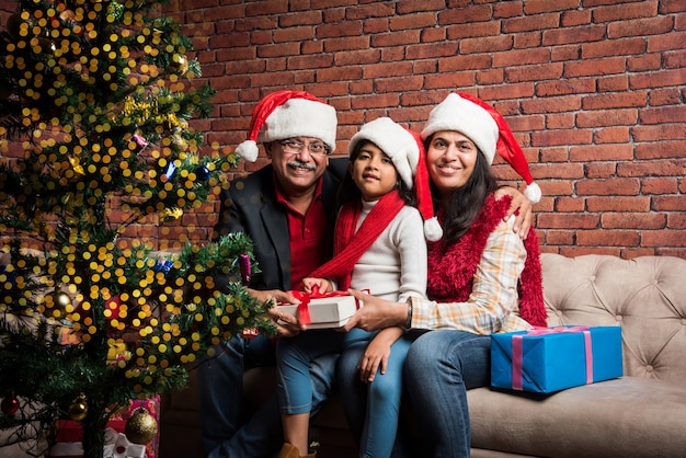 Petite fille asiatique indienne mignonne célébrant Noël avec les grands-parents et l'arbre de Noël alors qu'elle était assise sur un canapé et portant un bonnet de Noel et avec beaucoup de cadeaux