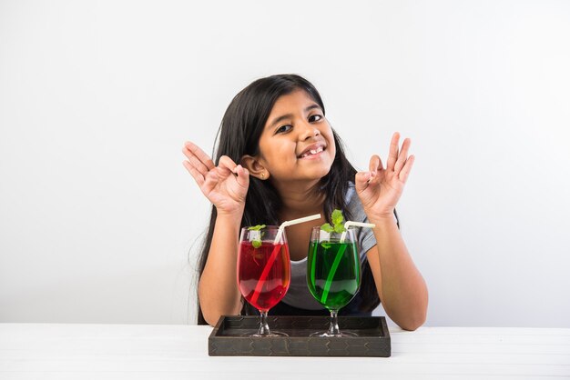 Petite fille asiatique indienne mignonne posant avec la boisson froide ou le jus frais à la table au-dessus du fond blanc