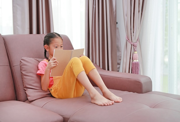 Petite fille asiatique lisant un livre allongé sur le canapé à la maison