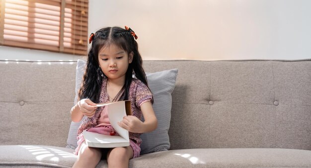 Une petite fille asiatique lit un livre sur un canapé confortable à la maison.