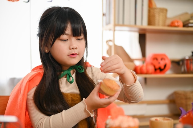Petite fille asiatique ludique en costume d'Halloween tenant un cupcake décorant son cupcake