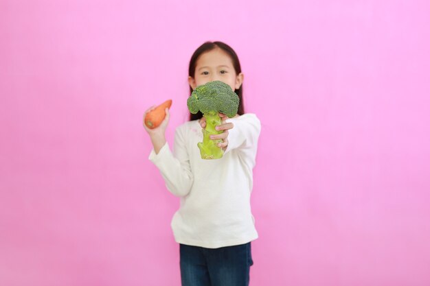 Petite fille asiatique montrant le brocoli debout isolé sur fond rose. Concept d'enfant et de légume. Focus sur le brocoli à la main