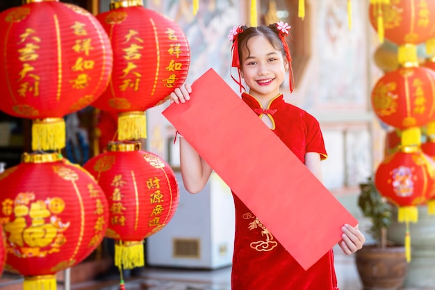 Petite fille asiatique portant un cheongsam chinois traditionnel rouge, montrer du papier blanc rouge et des lanternes avec le texte chinois Les bénédictions écrites dessus est une bénédiction Fortune pour le Nouvel An chinois