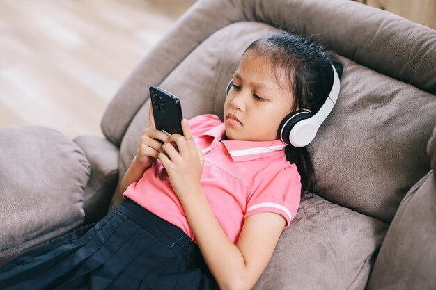 Une petite fille asiatique portant des écouteurs sans fil assise avec un téléphone portable à la maison