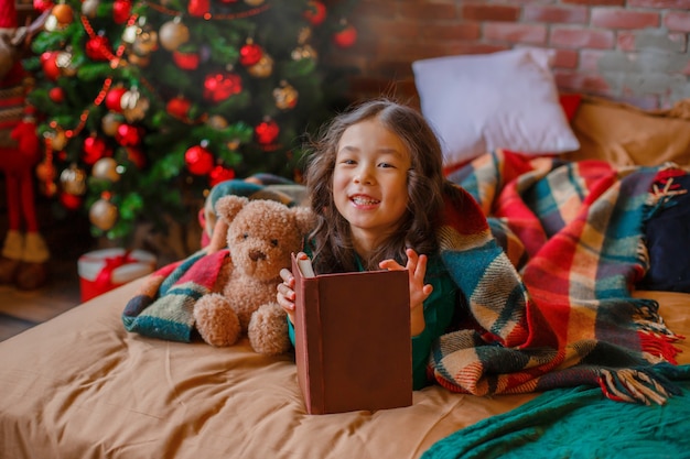 Petite fille asiatique en pyjama dans la chambre allongée sur le lit près de l'arbre de Noël en lisant un livre