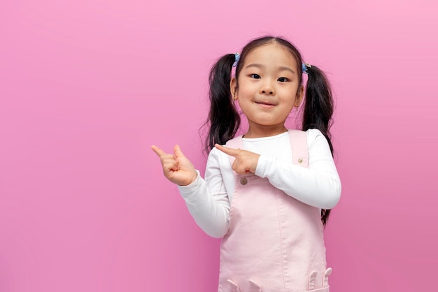 Petite fille asiatique en robe d'été rose aux cheveux longs montre ses mains sur le côté sur fond rose