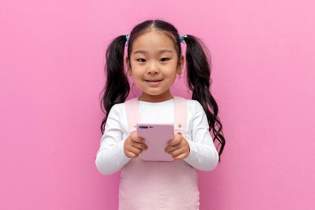 petite fille asiatique en robe d'été rose utilise un smartphone et choisit en ligne sur fond rose isolé