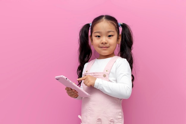 petite fille asiatique en robe d'été rose utilise un smartphone et choisit en ligne sur fond rose isolé