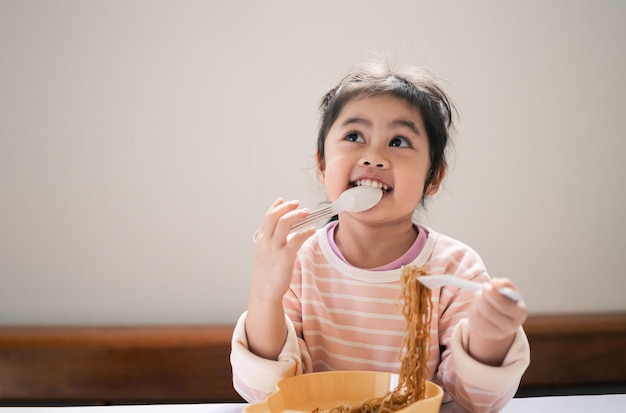 Une petite fille asiatique s'amuse avec une cuillère à couverts et une fourchette en mangeant de délicieuses nouilles dans la cuisine sur la table à manger