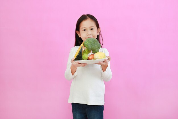 Petite fille asiatique souriante tenant une assiette de légumes frais en se tenant debout sur fond rose.