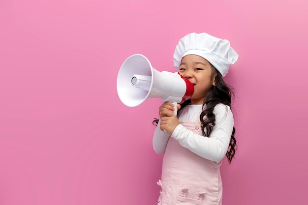 Petite fille asiatique en uniforme de chef annonce des nouvelles et des informations avec un mégaphone sur fond rose