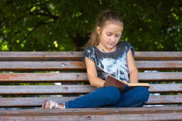 Petite fille assise sur un banc dans le parc et lisant.