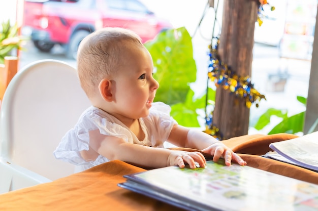 Petite fille assise sur une chaise haute pour bébé dans un café de la rue par table Enfants en robe blanche parlant