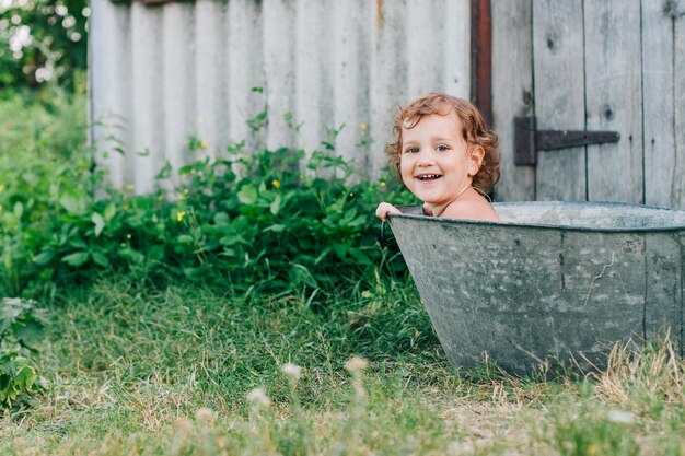 Photo petite fille assise dans une baignoire en métal dans un jardin verdoyant en été chaud sourire à pleines dents regarder la caméra