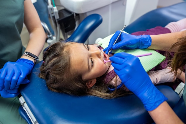 Petite fille assise dans un fauteuil dentaire et deux femmes dentistes traitant les dents de lait Deux femmes dentistes divertissent un petit patient