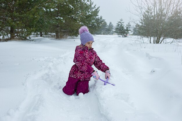 Une petite fille assise dans la neige fait des boules de neige à l'aide d'un outil de sculpture en plastique