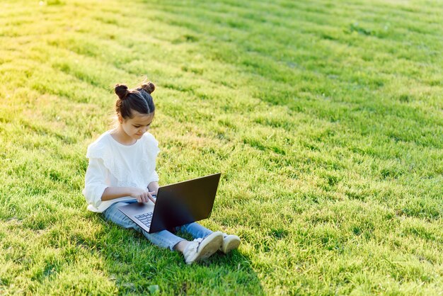 Petite fille assise dans le parc et travaillant avec un ordinateur portable. Éducation, mode de vie, concept technologique