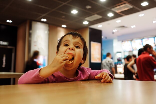 Petite fille assise seule au tabel et mange un repas au café ou au restaurant. Mettez le repas dans la bouche ouverte.