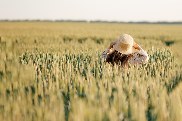 La petite fille attirante dans un chapeau de paille court sur un champ de blé avec les oreilles vertes