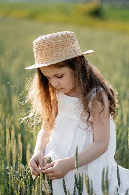 La petite fille attirante dans un chapeau de paille court sur un champ de blé avec les oreilles vertes